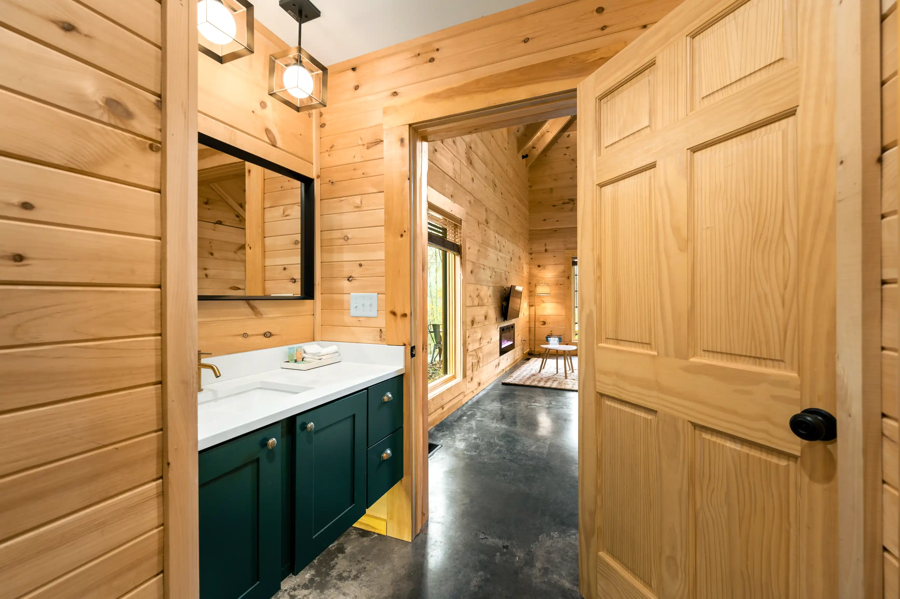 Rustic wooden interior view of a cabin showing a bathroom with a teal vanity and a room with a view of a living space through an open door.