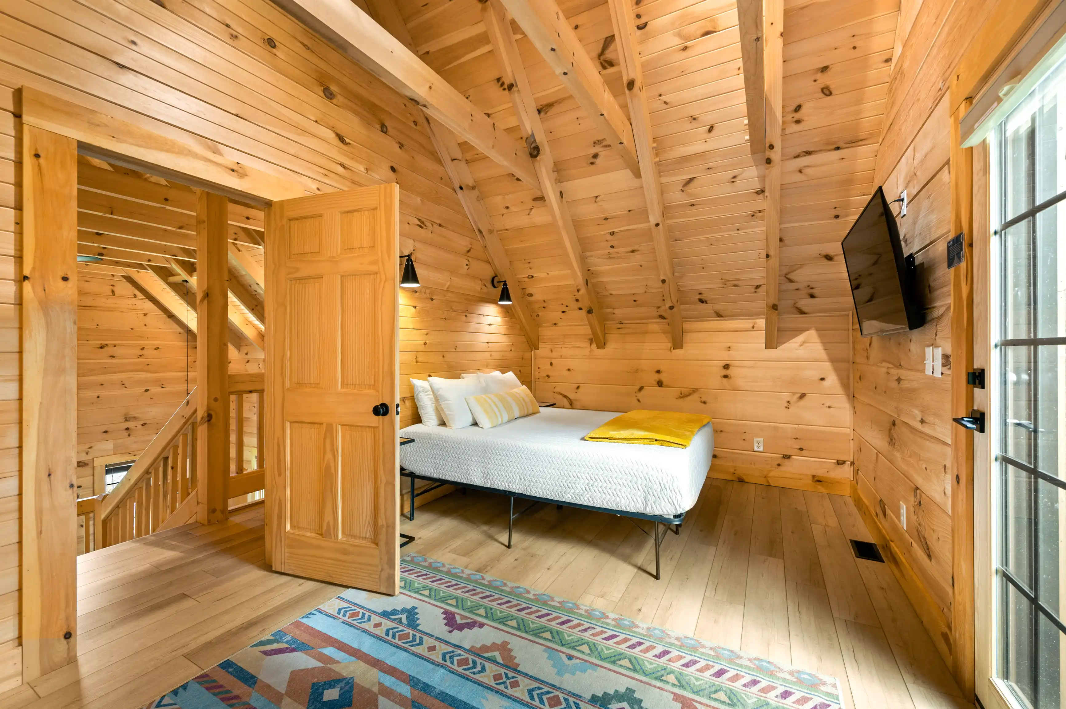 Cozy wooden cabin bedroom with a large bed, colorful rug, vaulted ceiling, and natural light from adjacent windows.