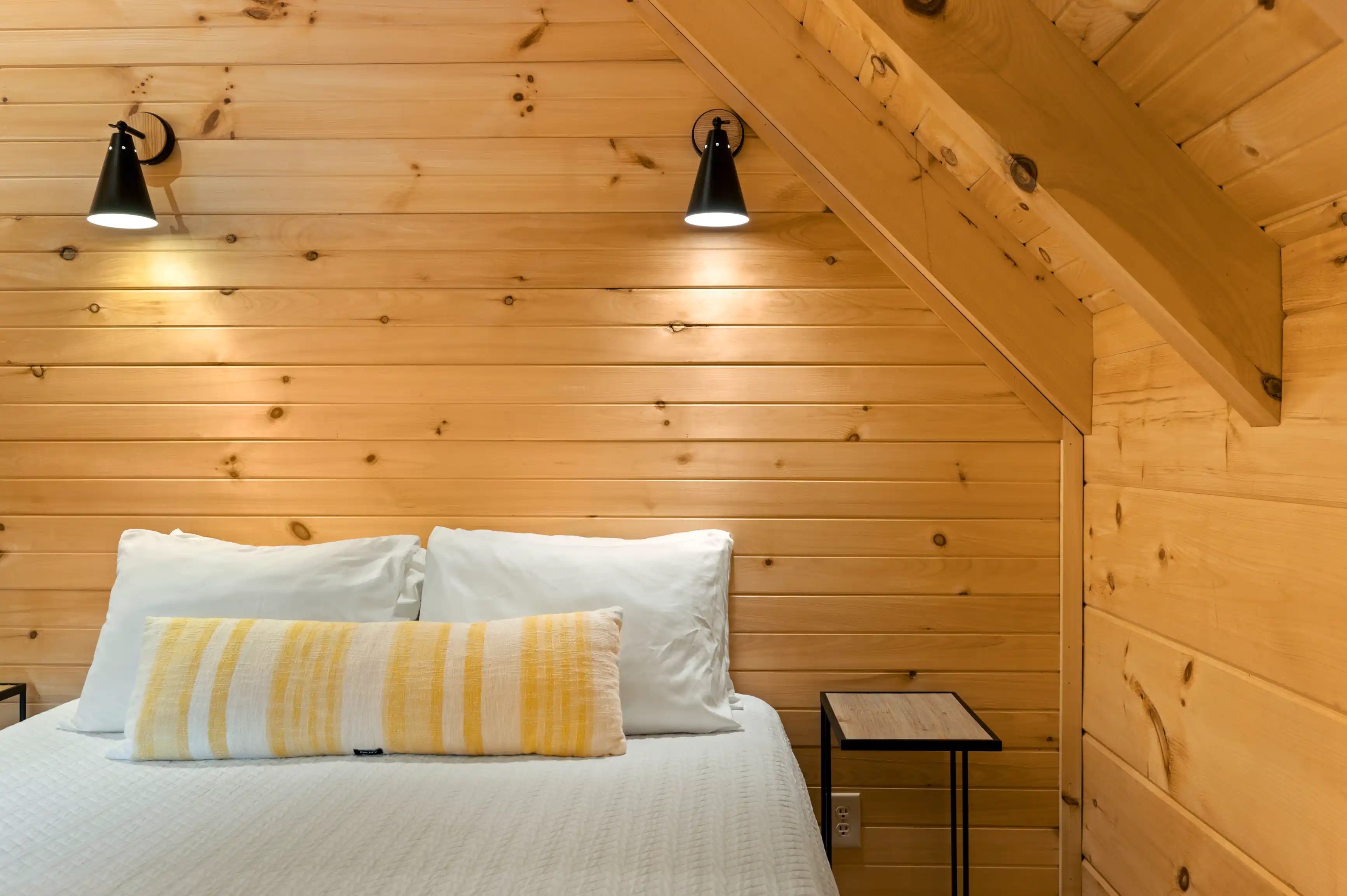 Cozy bedroom interior with white bedding, a yellow striped pillow, wooden walls, and wall-mounted lights.