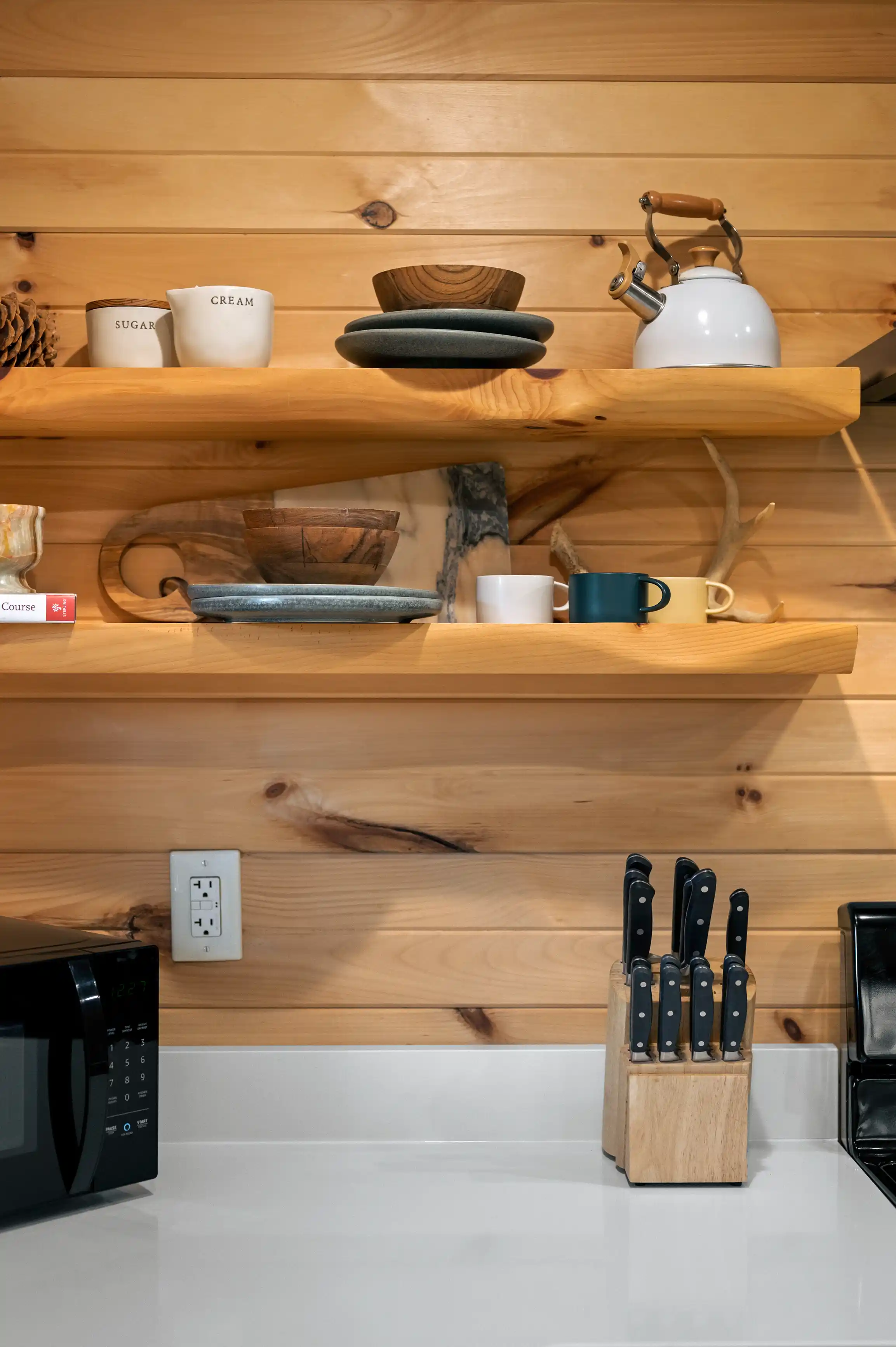 A cozy kitchen interior with wooden shelves holding a tea kettle, mugs, and canisters labeled 'SUGAR' and 'CREAM', above a counter with a knife block and a microwave.