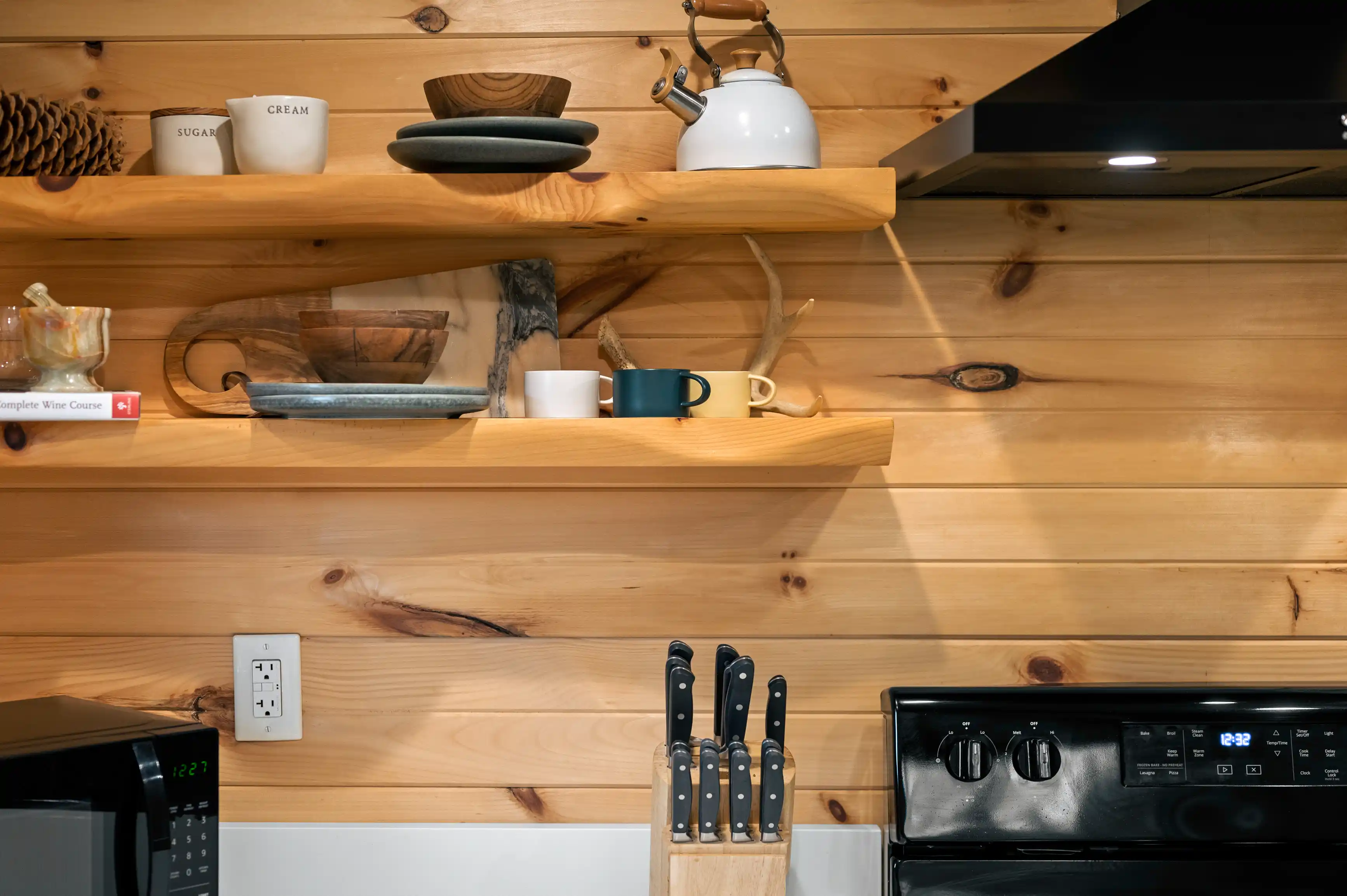 A cozy kitchen interior with wooden shelves displaying utensils, cups, and a kettle, above a stove and a set of knives to the side.