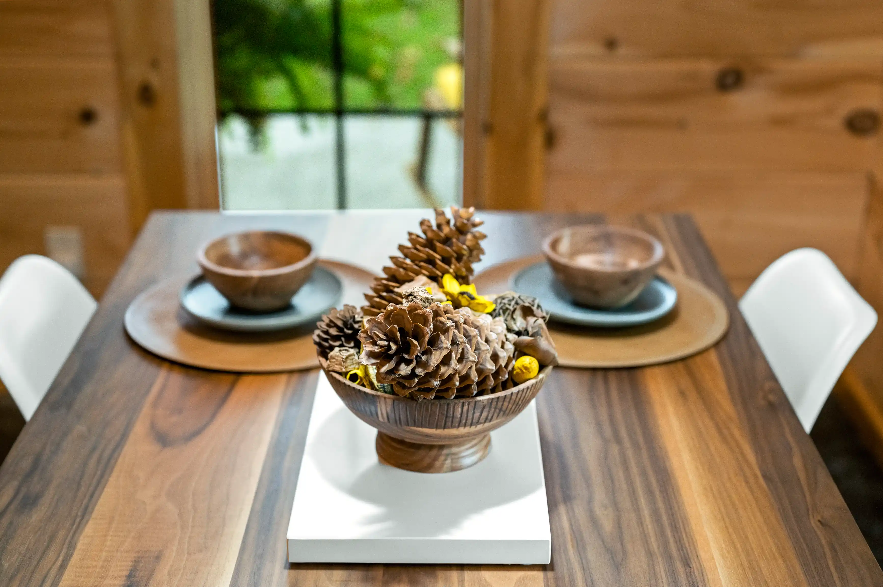 A wooden dining table set with a centerpiece of pine cones in a bowl, flanked by two wooden bowls on plates, with white chairs and a window view in the background.