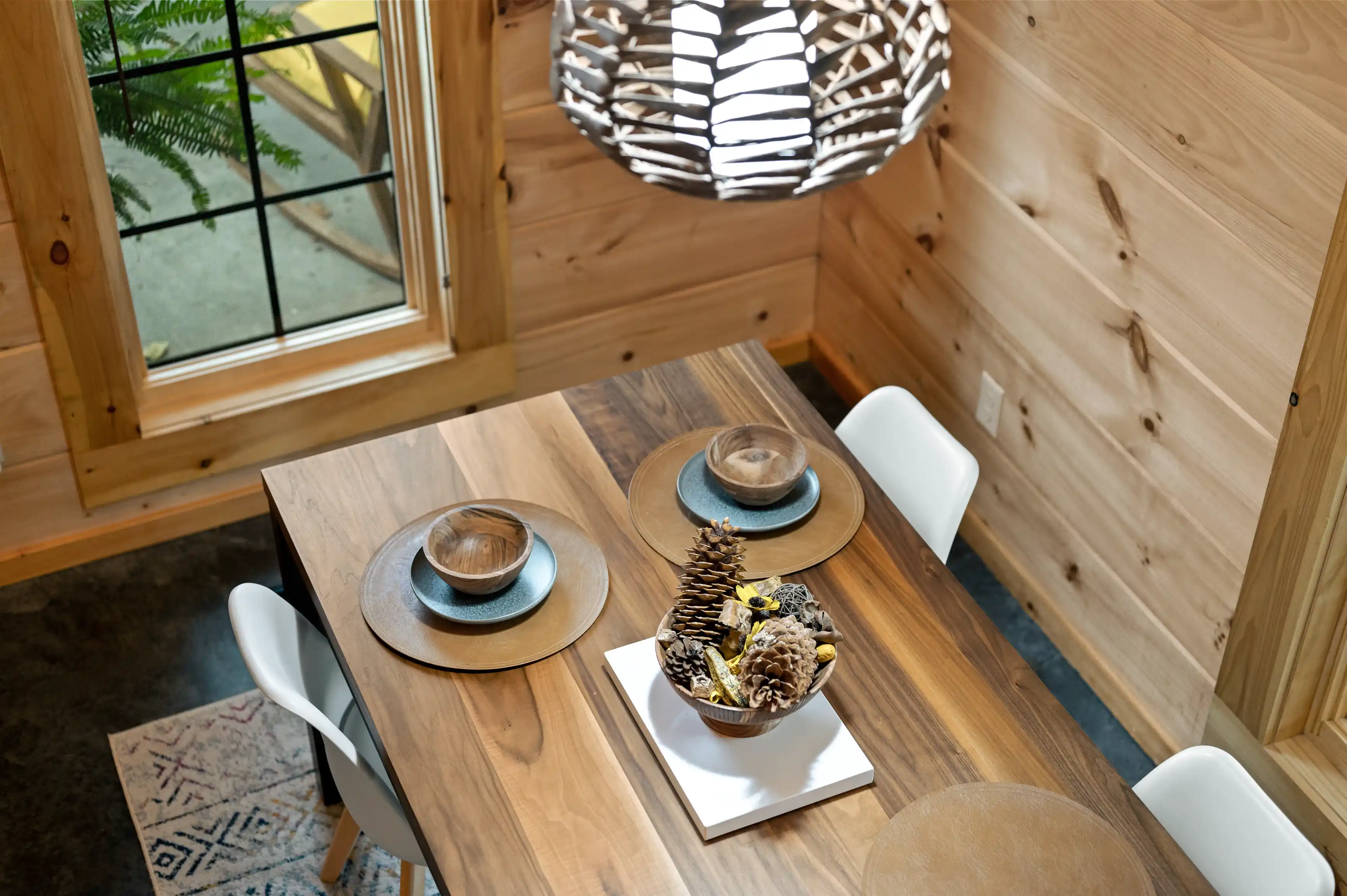 Cozy wooden interior of a dining area with modern furnishings, a decorative centerpiece on the table, and a unique pendant light above, near a window overlooking greenery.