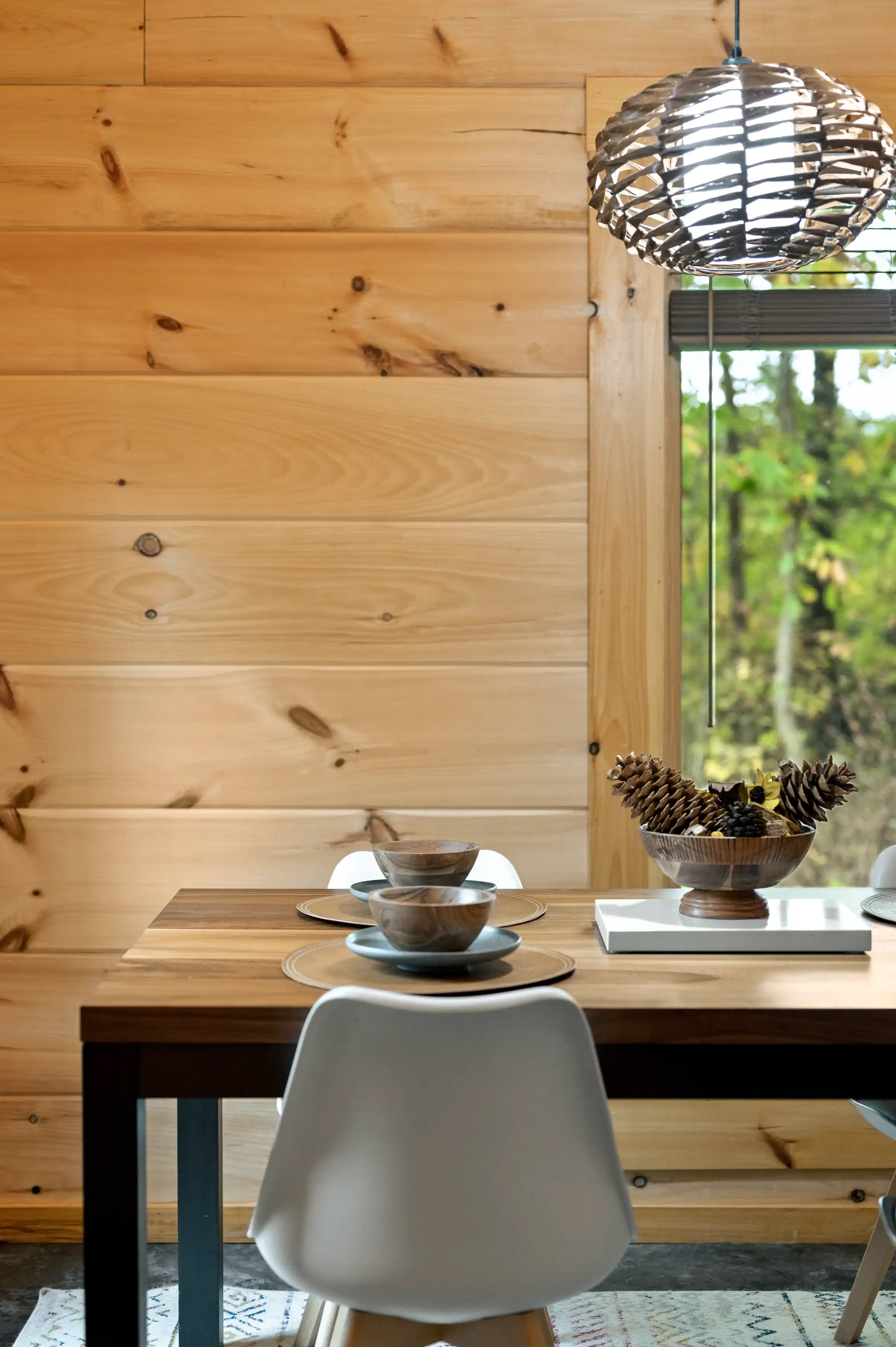 Contemporary dining setup with a wooden table, modern white chairs, a decorative bowl of pine cones, and a stylish pendant lamp in a room with wooden walls and a view of trees through the window.