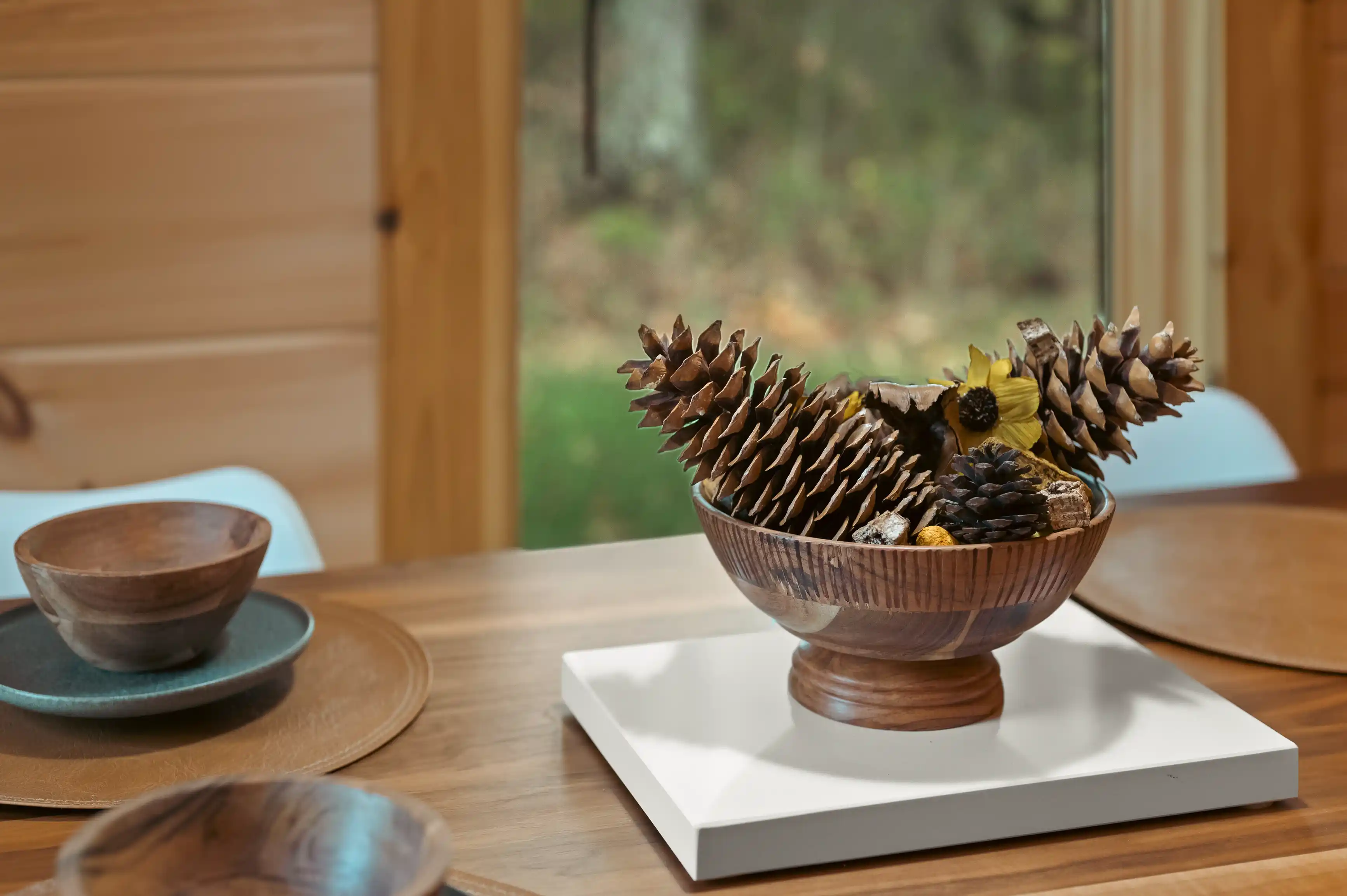Wooden bowl filled with pine cones on a white tray on a wooden table, with other wooden dishes and a blurred forest view in the background.