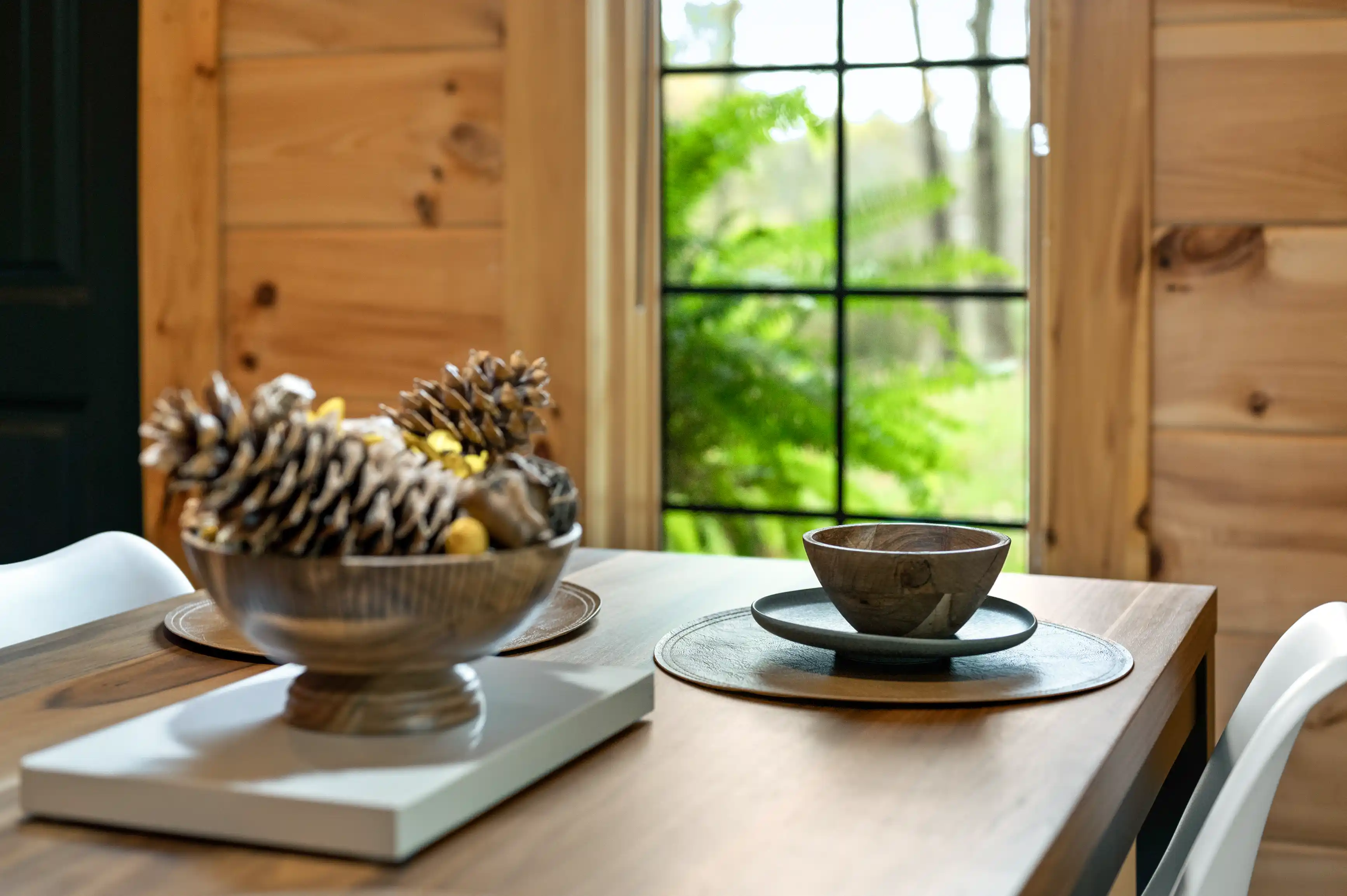 A cozy dining area with a wooden table set with a bowl and plate, a centerpiece of pine cones, and a view of greenery through a windowpane.