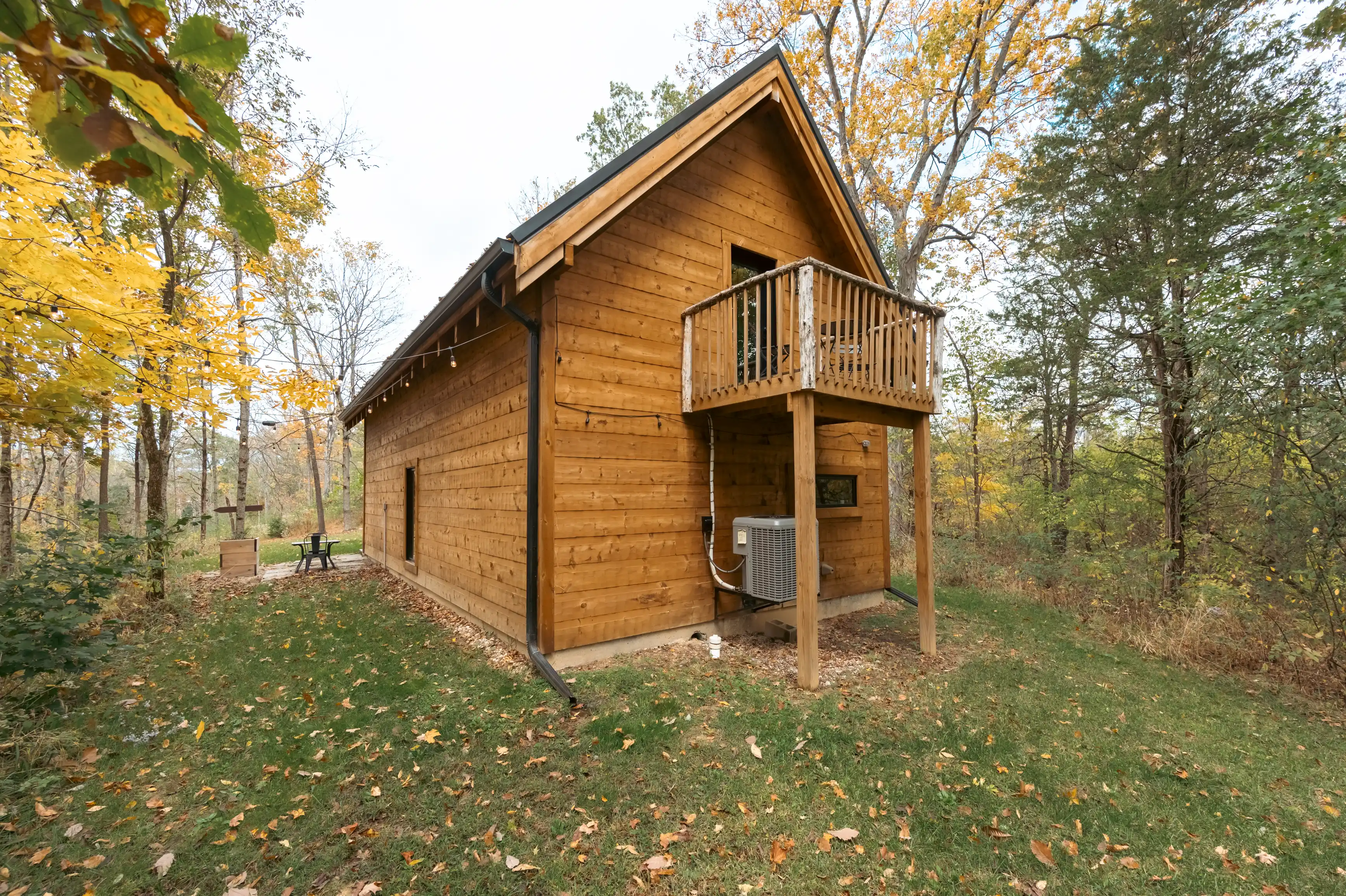 A wooden cabin with a balcony amidst autumn foliage, with a picnic table and chairs on the lush green grass.