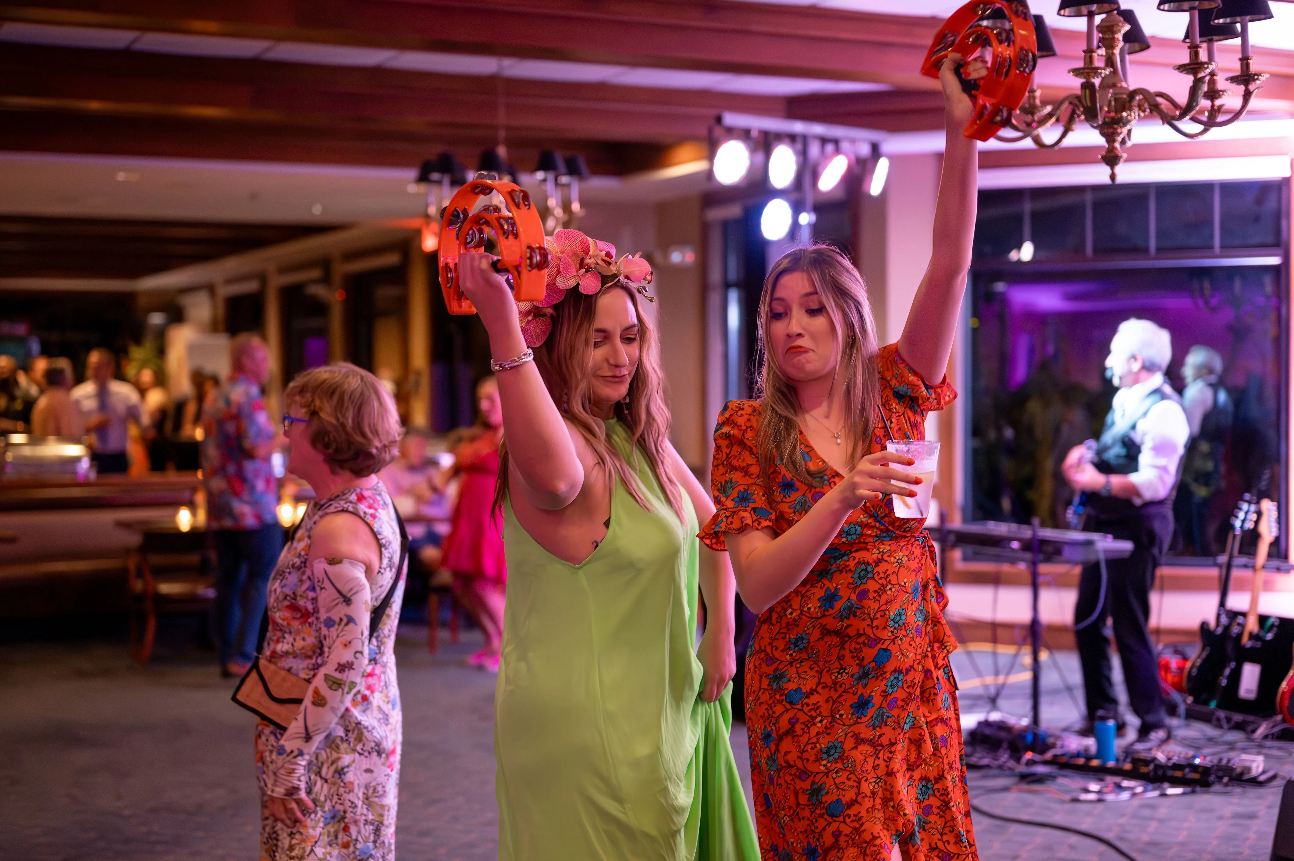 Two women dancing holding flower bouquets at a festive event with a live band in the background.