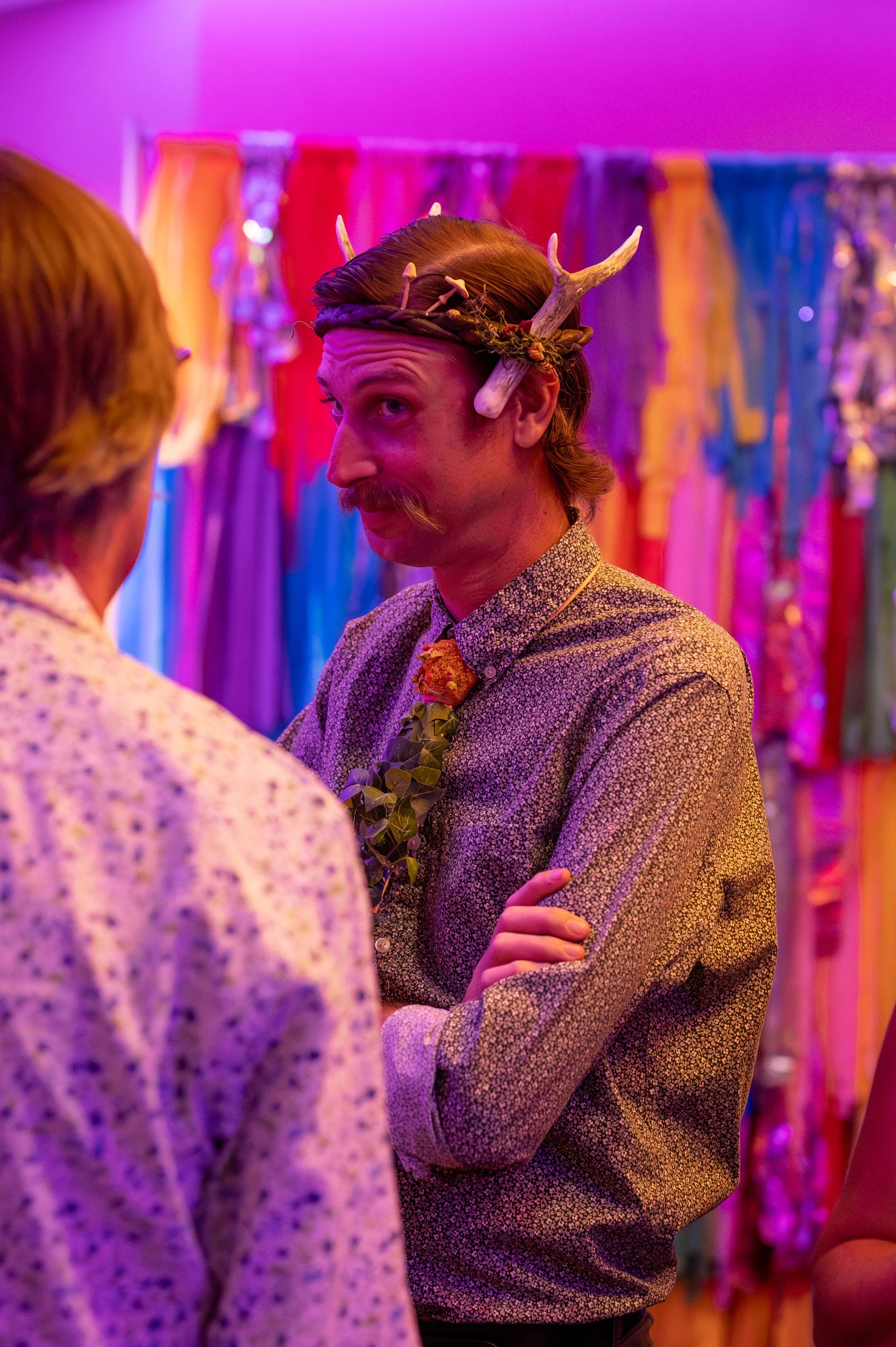 Person wearing a headband with horns talking to another person in a brightly lit purple room with colorful curtains in the background.
