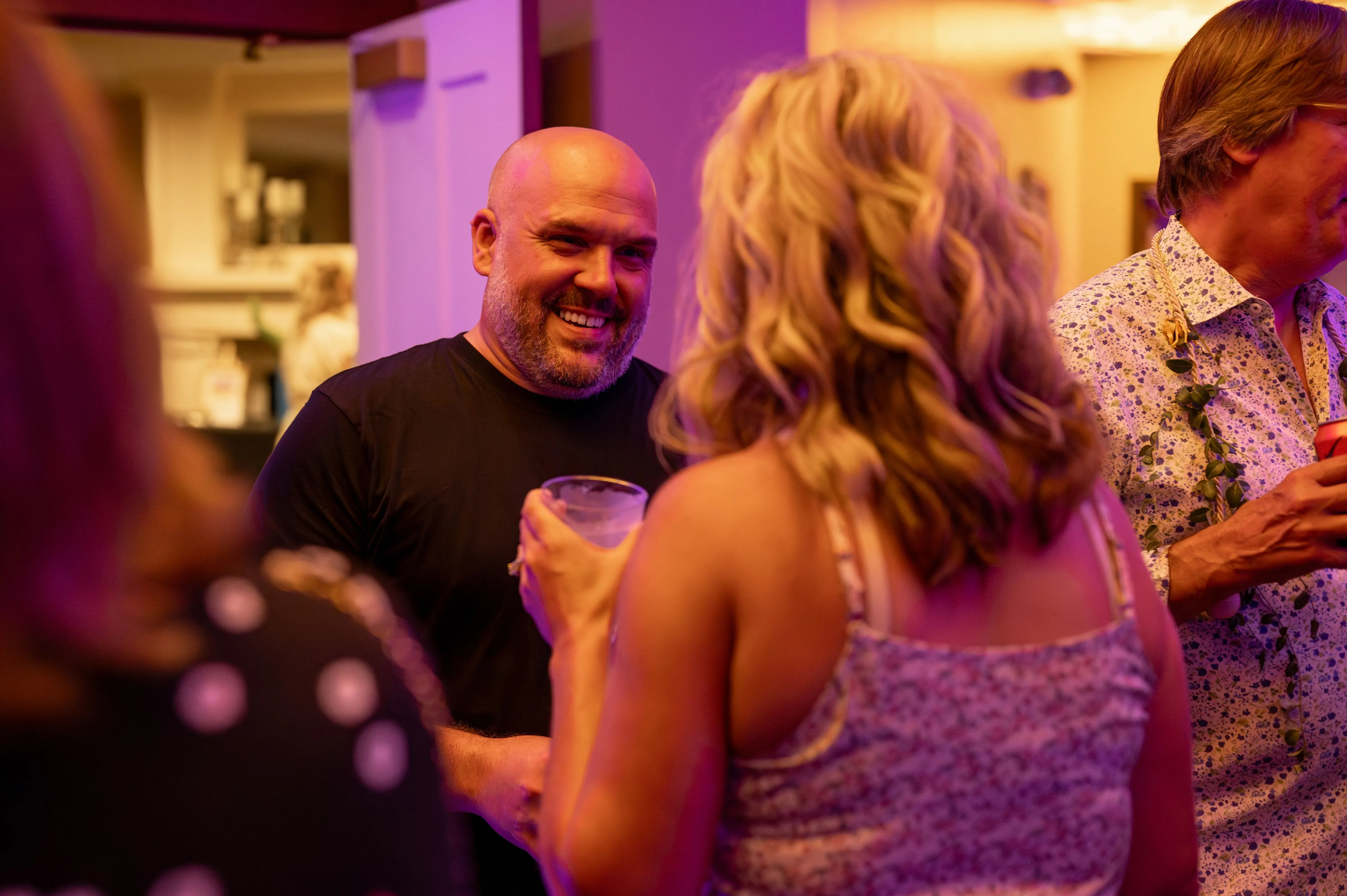 People socializing at an indoor gathering, with a smiling man holding a drink in focus.