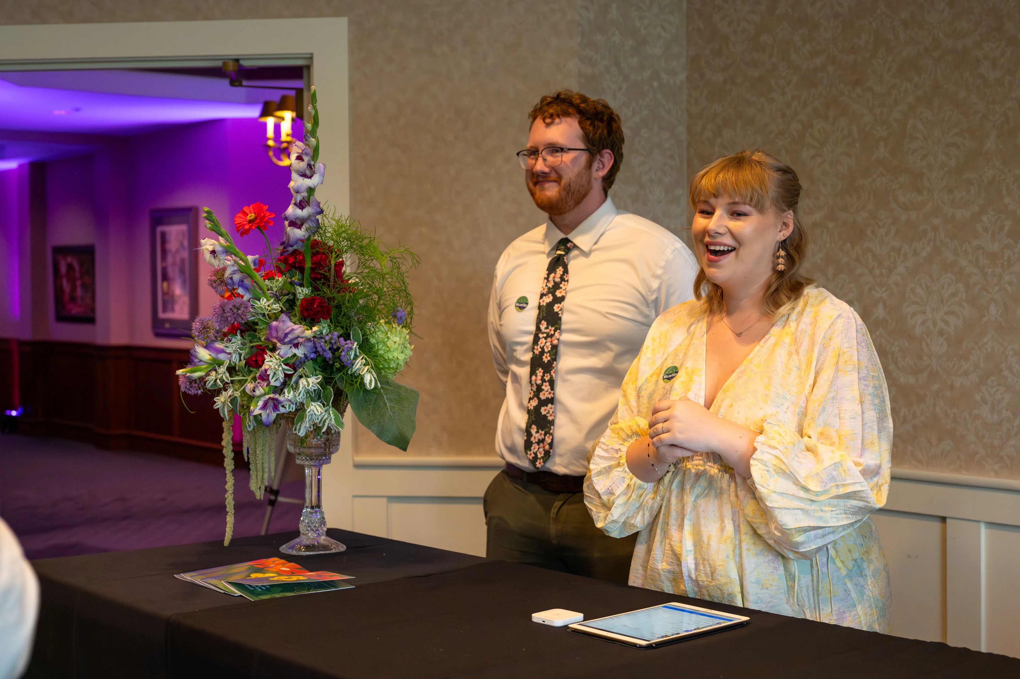 Two people standing at a table with a floral arrangement, smiling and clapping in a room with purple lighting.