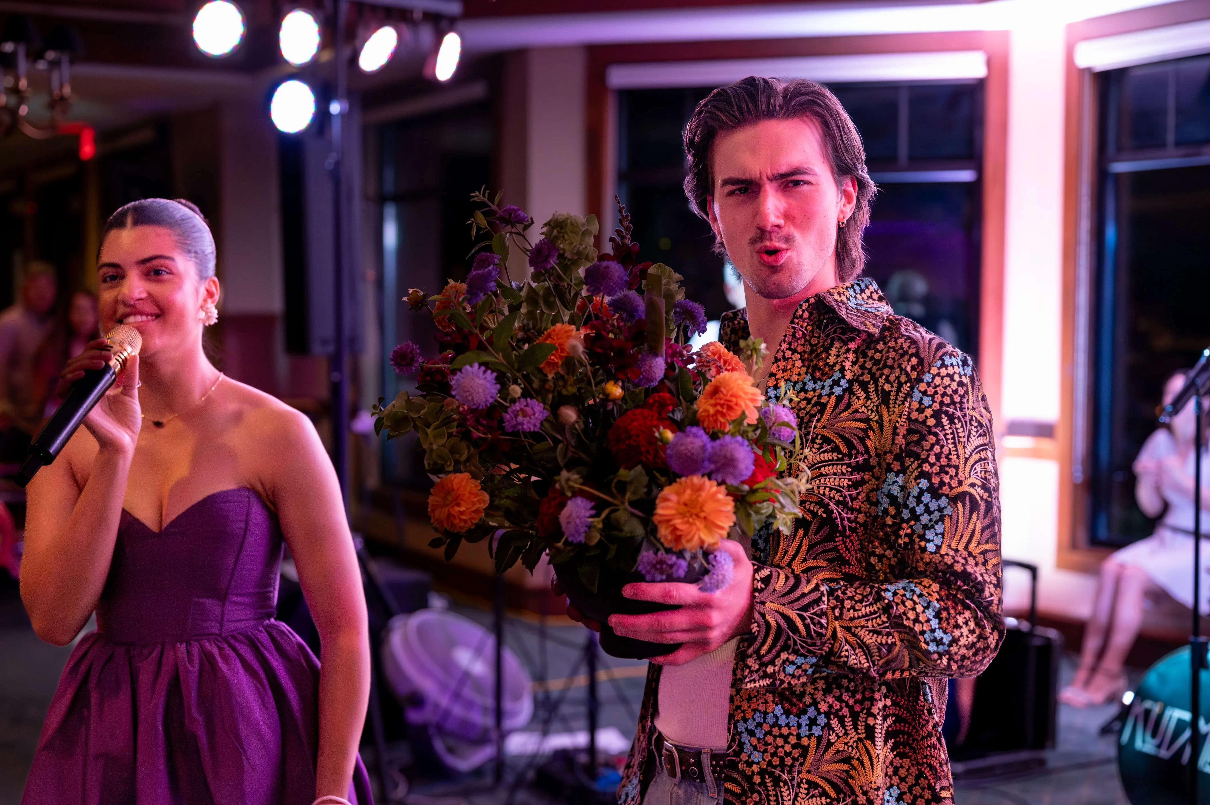 Two people at a party, a woman in a purple dress singing into a microphone and a man in a patterned shirt holding a bouquet of flowers with a playful expression.