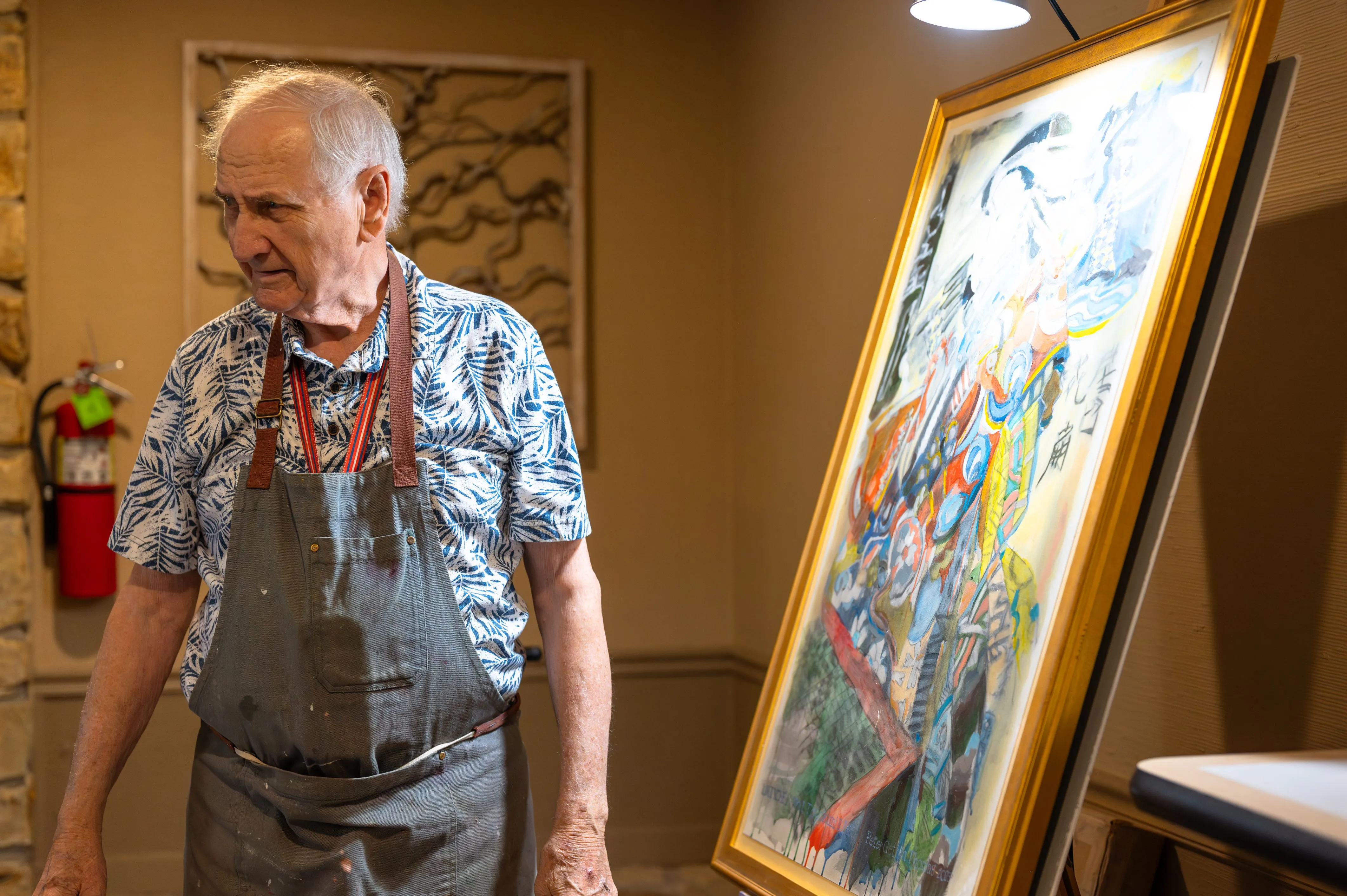 Elderly person in a patterned shirt and pants standing beside a framed colorful painting in a room with patterned wallpaper.
