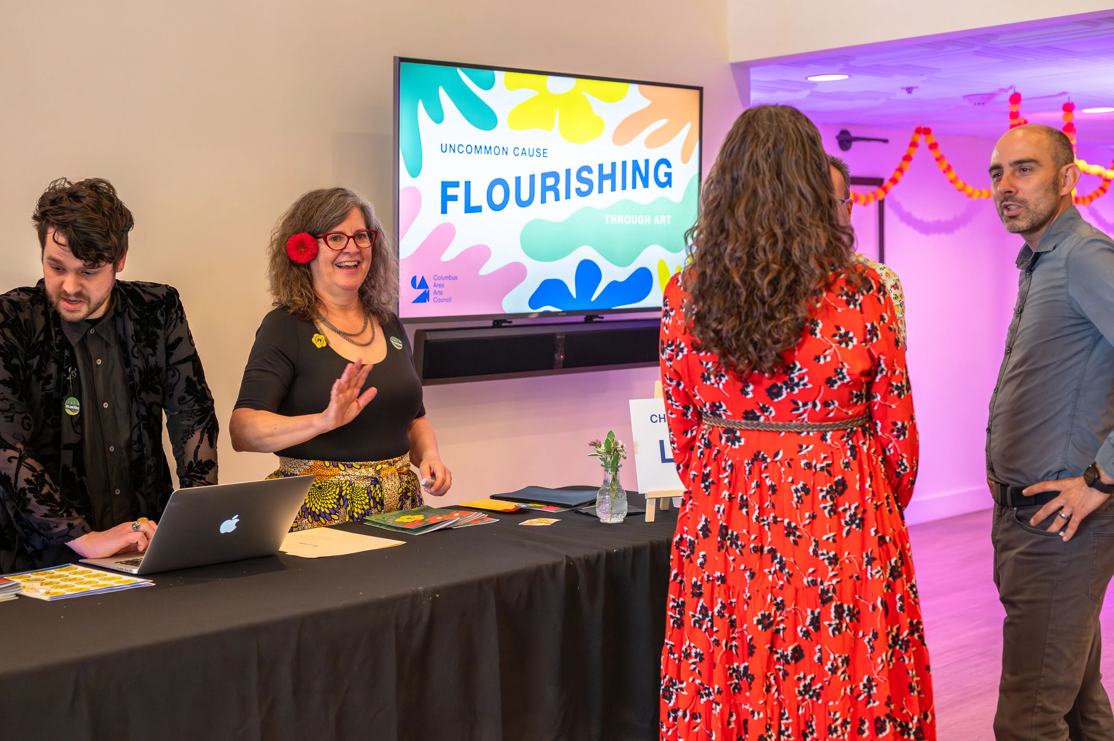 People interacting at a conference registration desk with a "FLOURISHING" sign displayed on a screen in the background.