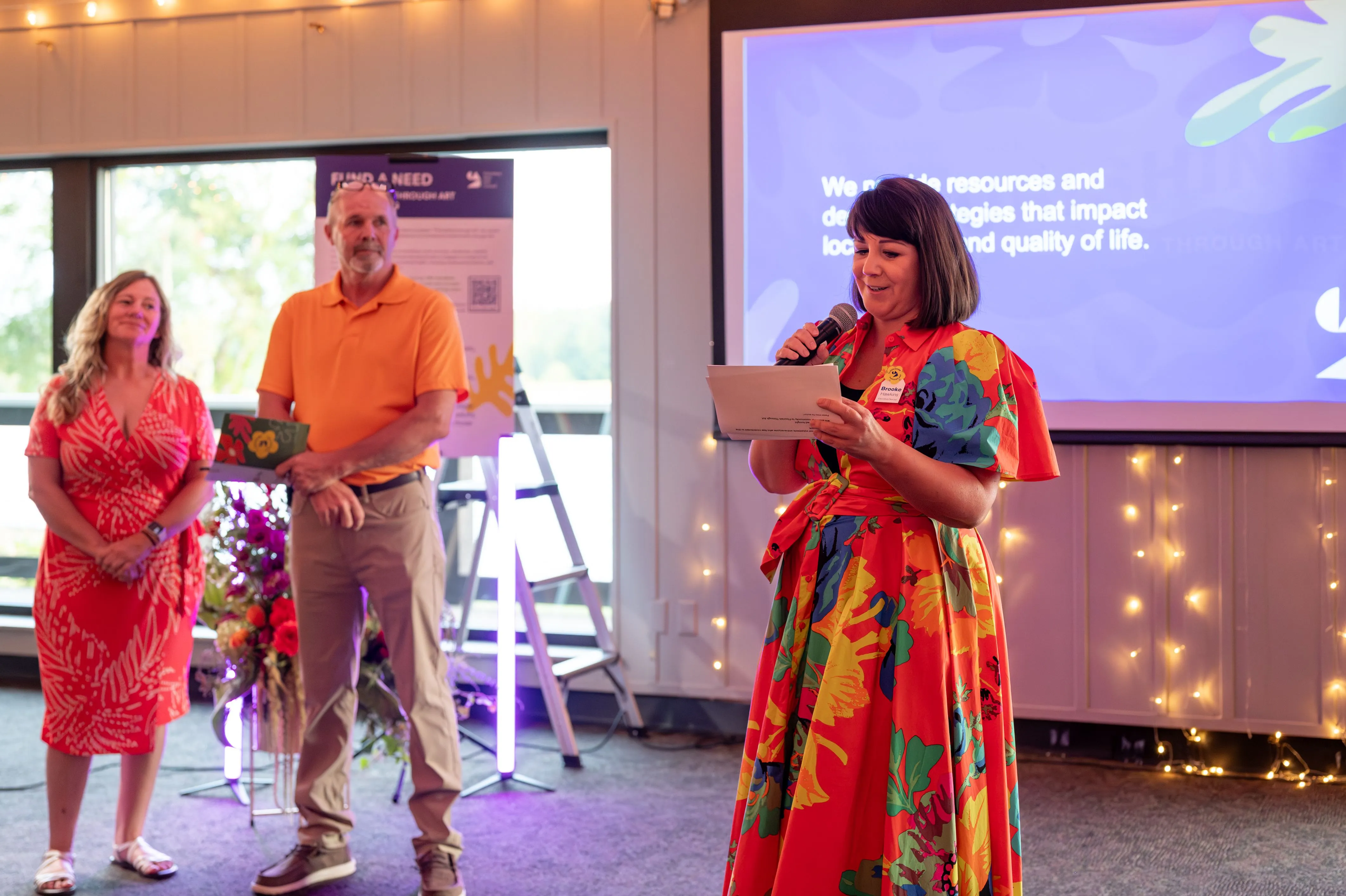Three people at an indoor event with one of them reading from a paper and a projection screen in the background displaying text and a floral graphic.
