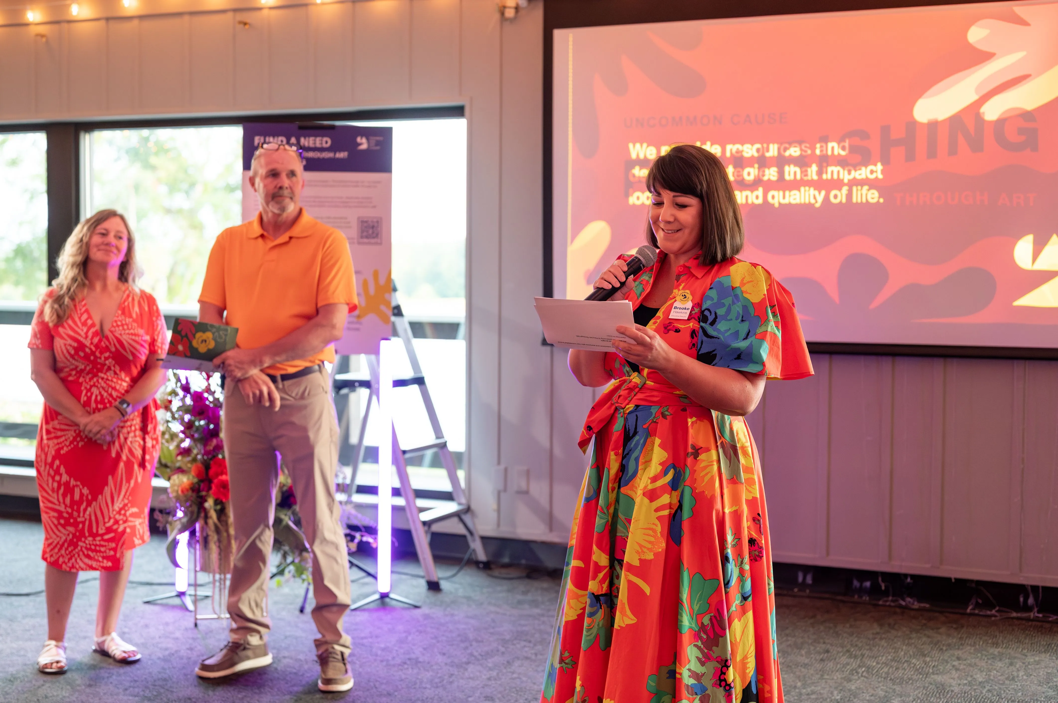 A woman in a colorful dress speaking into a microphone at an event with two other individuals behind her, one carrying flowers, against a backdrop with a projected image.