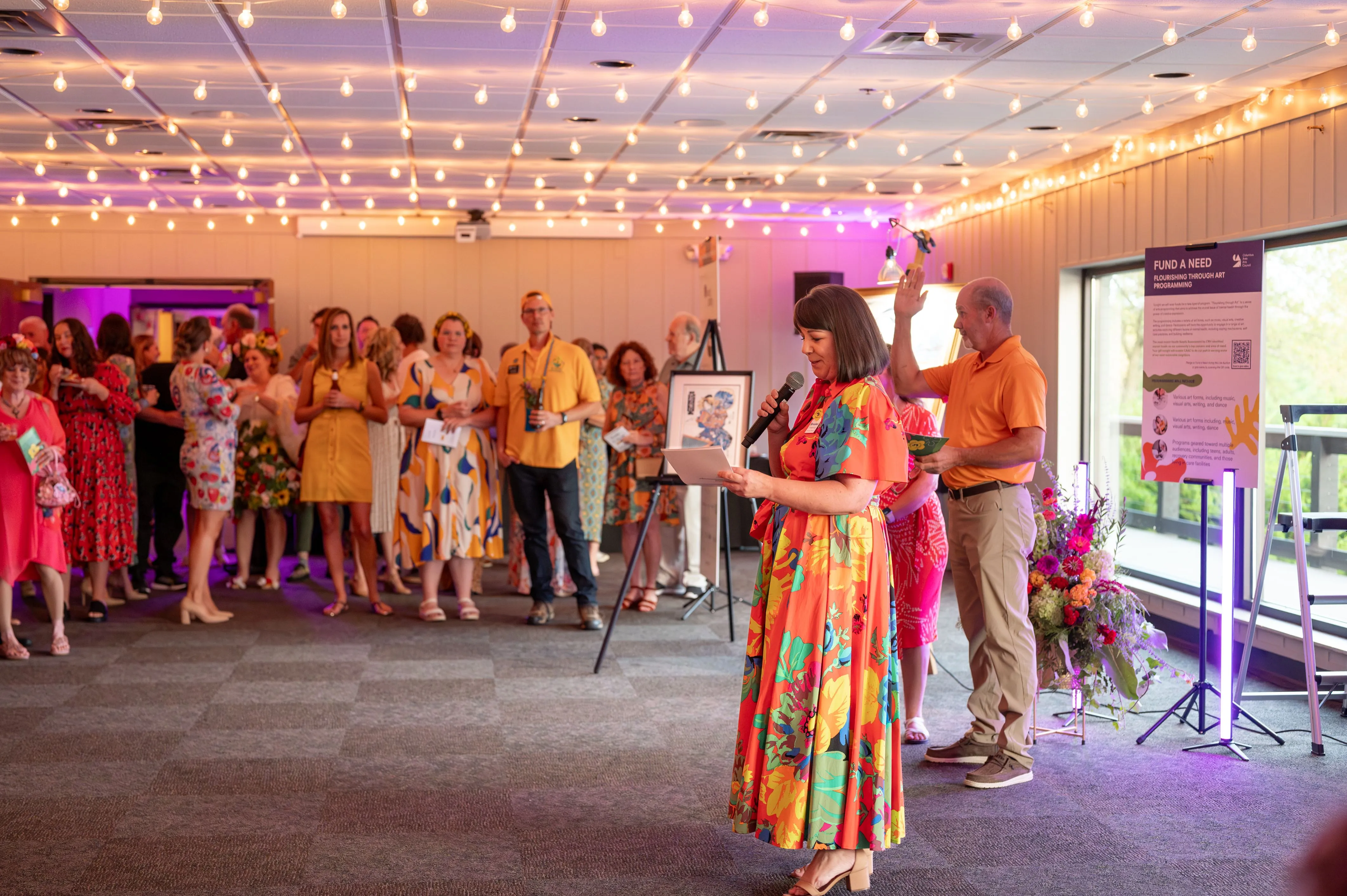 A group of people gathered at an indoor event with a woman in a colorful dress holding a framed picture in the foreground.