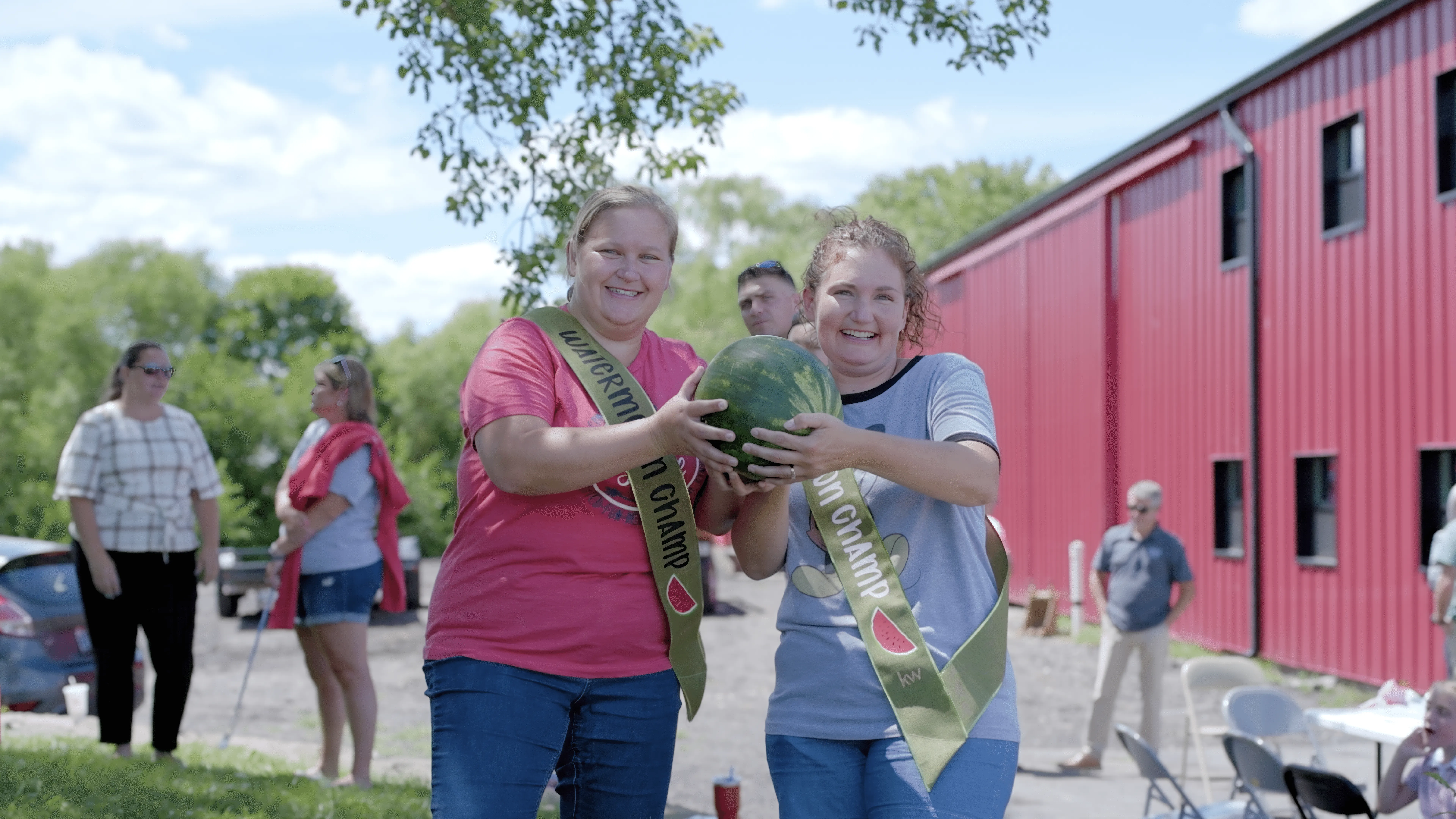 Two people holding a large watermelon with smiles, at an outdoor event with others in the background.