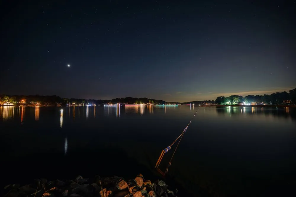Nighttime view of a calm lake with sparkling stars overhead and reflections of nearby lights on the water's surface.
