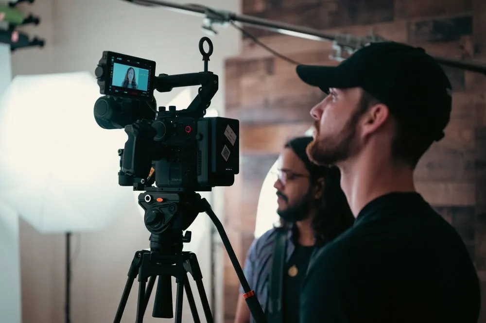 Professional video camera on a tripod with a monitor displaying a woman, and two male crew members in the background monitoring the shoot.