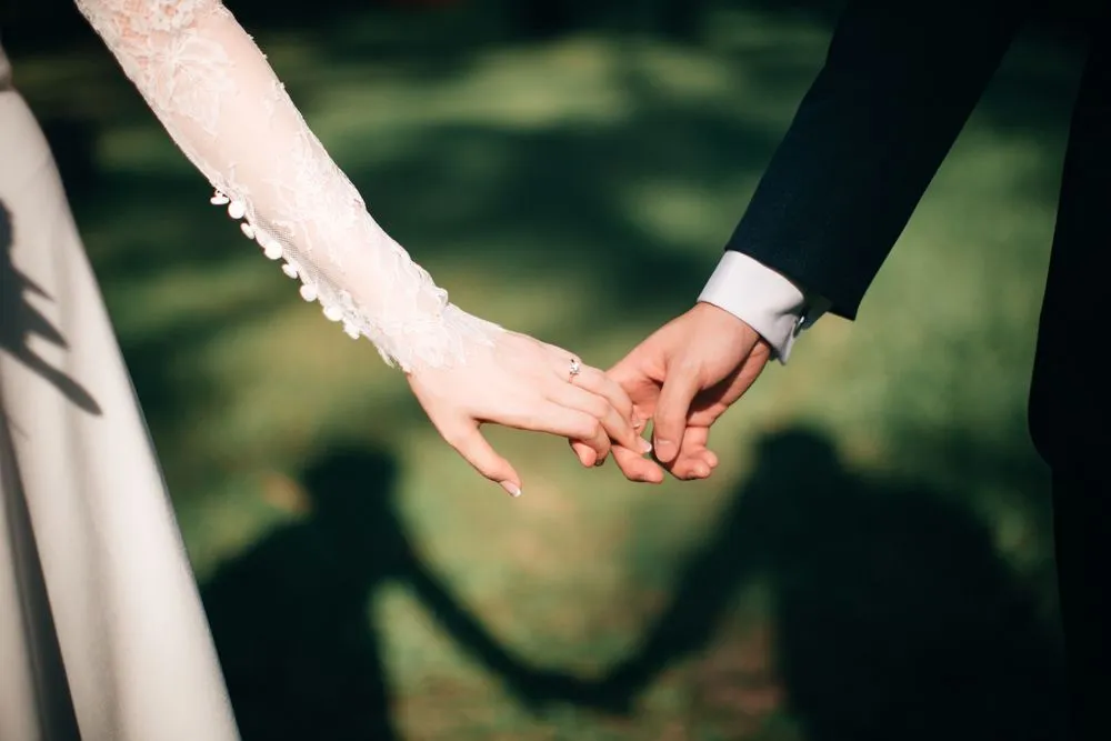 Close-up of a bride and groom holding hands, with their shadows visible on the grass.