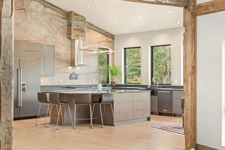 Modern kitchen interior with island, bar stools, and stainless steel appliances, featuring wooden beams and stone wall accents with a view of trees through large windows.