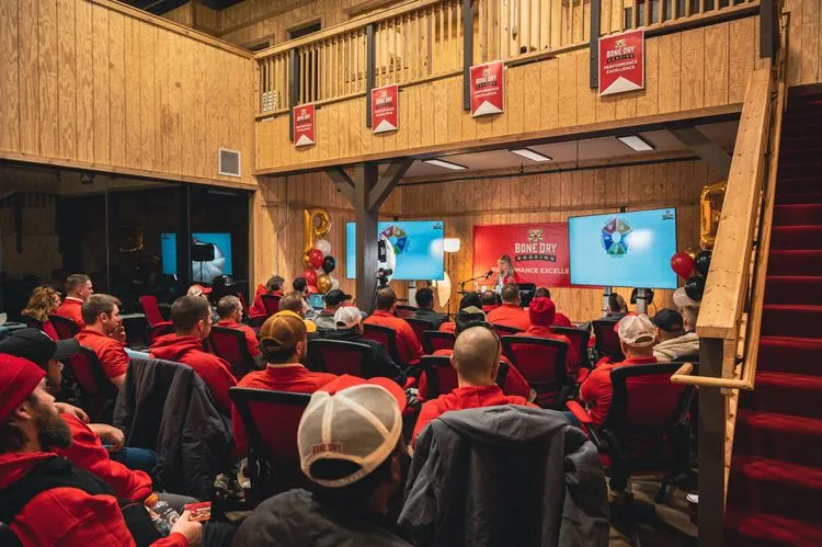 Group of people in red attire attending a presentation in a room with wooden interiors and "Bone Dry Roofing" banners.