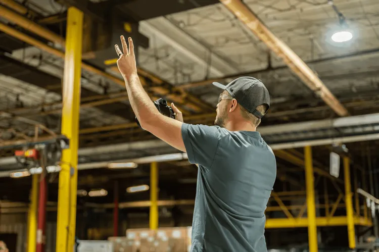 Man in a warehouse wearing a baseball cap, gesturing with his hand in the air, holding a walkie-talkie, with industrial shelving in the background.