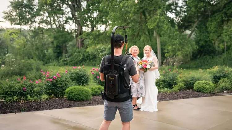 Photographer with a backpack and camera gear facing two smiling women in bridal attire in a garden with pink flowers and greenery in the background.