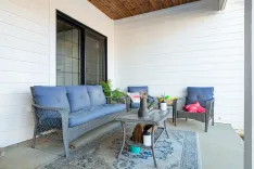 A cozy outdoor patio area with a wicker sofa and chair set featuring blue cushions, a coffee table with decorative items, and green plants, against a white siding house with sliding glass doors.