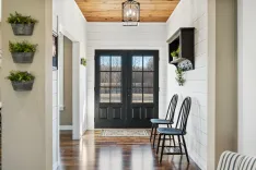 Bright entryway with double black doors, wooden ceiling, wall-mounted planters, and black chairs.