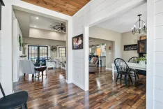 Spacious interior of a modern home with a wooden ceiling, hardwood floors, white walls, and a view into the living room with a comfortable seating arrangement.