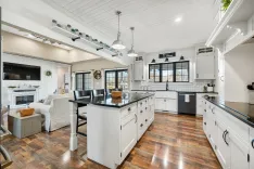 A spacious, well-lit kitchen with white cabinetry, black countertops, and hardwood floors, featuring an island, stainless steel appliances, and a seating area with a fireplace.