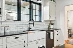 Modern farmhouse-style kitchen with white shaker cabinets, black countertops, a white subway tile backsplash, and a large apron-front sink beneath black-framed windows.