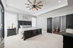 Elegant master bedroom with black furniture, white bedding, a ceiling fan, and an en-suite bathroom visible in the background.