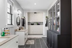 Elegant bathroom interior with dark cabinetry, subway tiles with decorative border, dual sinks with mirrors, and a walk-in shower.