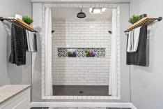 A modern bathroom with a walk-in shower featuring white subway tiles, a decorative tile accent, two black and white striped towels hanging on the side, and a plant on a shelf.