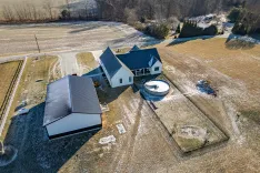 Aerial view of a rural residential property with a house, detached garage, above-ground pool, and surrounding fields in a sparse snowy landscape.