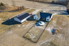 Aerial view of a rural residential property with a large house and detached garage, a circular driveway, and surrounding fields in winter, with sparse snow cover.