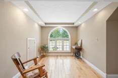 Bright room with beige walls, wooden floor, vaulted ceiling with recessed lighting, and a large arched window with a view of trees. A rocking chair, side table, and decorative plants are also visible.