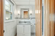 Modern bathroom interior with white cabinets, gray countertop, framed mirror, sconce lights, and a window letting in natural light.
