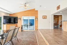 Spacious living room interior with brick tile flooring, wood accent wall with mounted flat-screen TV, ceiling fan, glass sliding door leading to a patio, and open doorway to another room. Decor includes bar-themed wall signs.