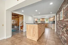 Spacious kitchen with wooden cabinets, tile floor, and brick wall accent, leading to a cozy living area with stairway.