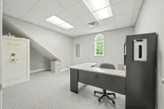Modern office interior with a large safe, L-shaped desk, rolling chair, filing cabinet, fluorescent ceiling lights, and a window showing greenery outside.