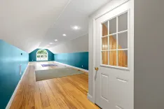 Spacious hallway with slanted ceilings, hardwood floors, teal walls, a white door with window panes, and a large arched window at the end allowing natural light.