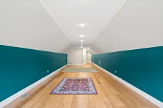 A long, narrow hallway with hardwood floors, white ceiling, and dark teal lower walls, featuring recessed lighting and a decorative red and blue rug.