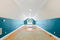 Empty spacious room with slanted white ceiling, teal walls, hardwood floor with a central area rug, and a large arched window at the end.