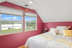 Bright bedroom with large windows overlooking a lake, red walls, white and yellow bedding, and a sloped ceiling.