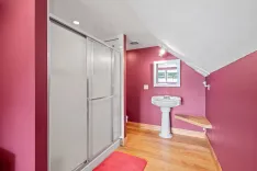 Interior of a bathroom with magenta walls, hardwood floors, a glass shower door, pedestal sink, and a small wooden shelf under a window with a view outside.