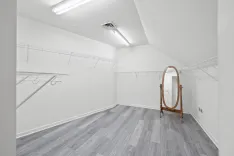 An empty walk-in closet with white walls, grey wood floor, built-in wire shelving, and a standalone wooden full-length mirror.